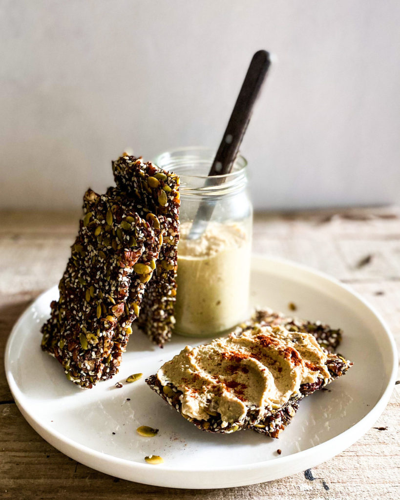 nuts and seeds crackers stacked against a jar of hummus and 1 cracker with hummus spread on it